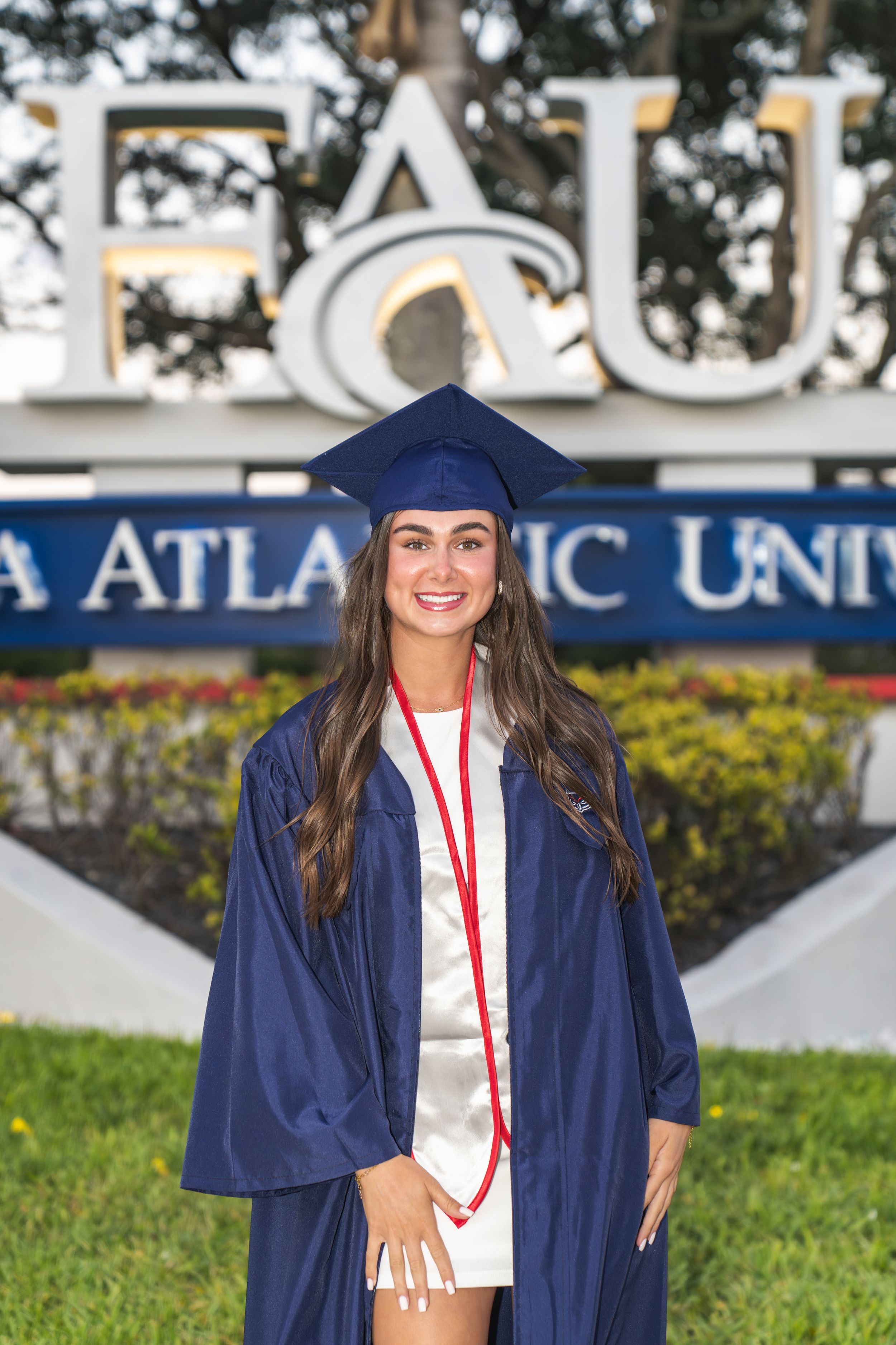 A smiling young woman in blue graduation cap and gown stands before Florida Atlantic University sign.