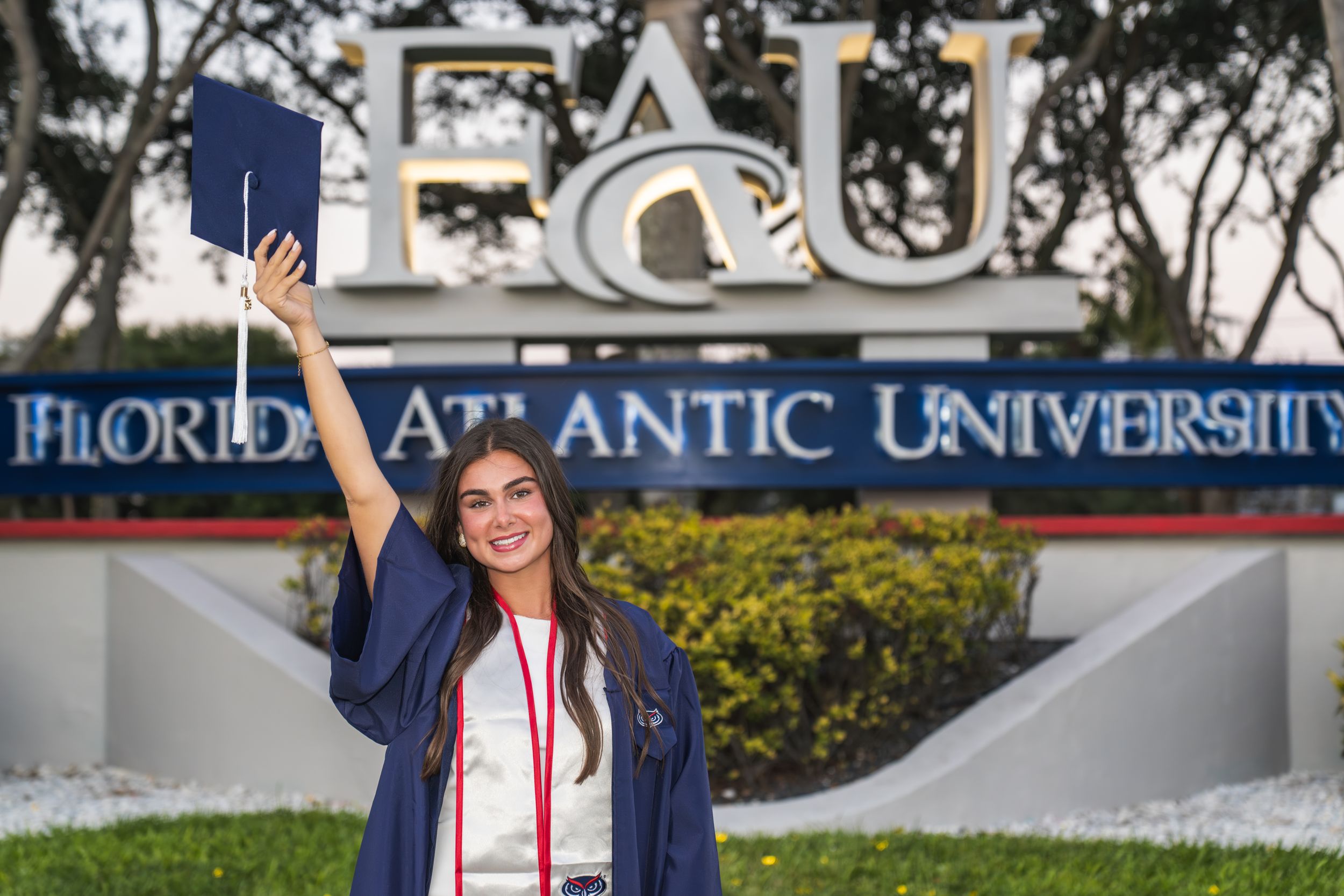 A smiling graduate in cap and gown raises her diploma in front of Florida Atlantic University sign.