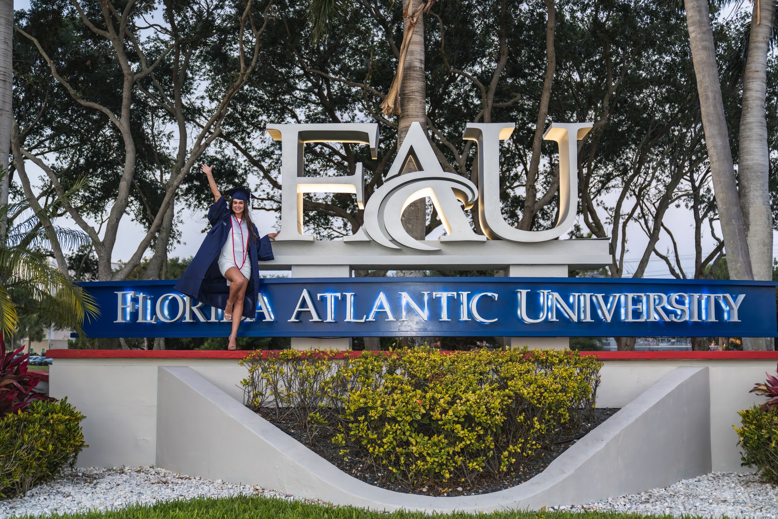 Graduate poses proudly in cap and gown at Florida Atlantic University FAU campus entrance sign.