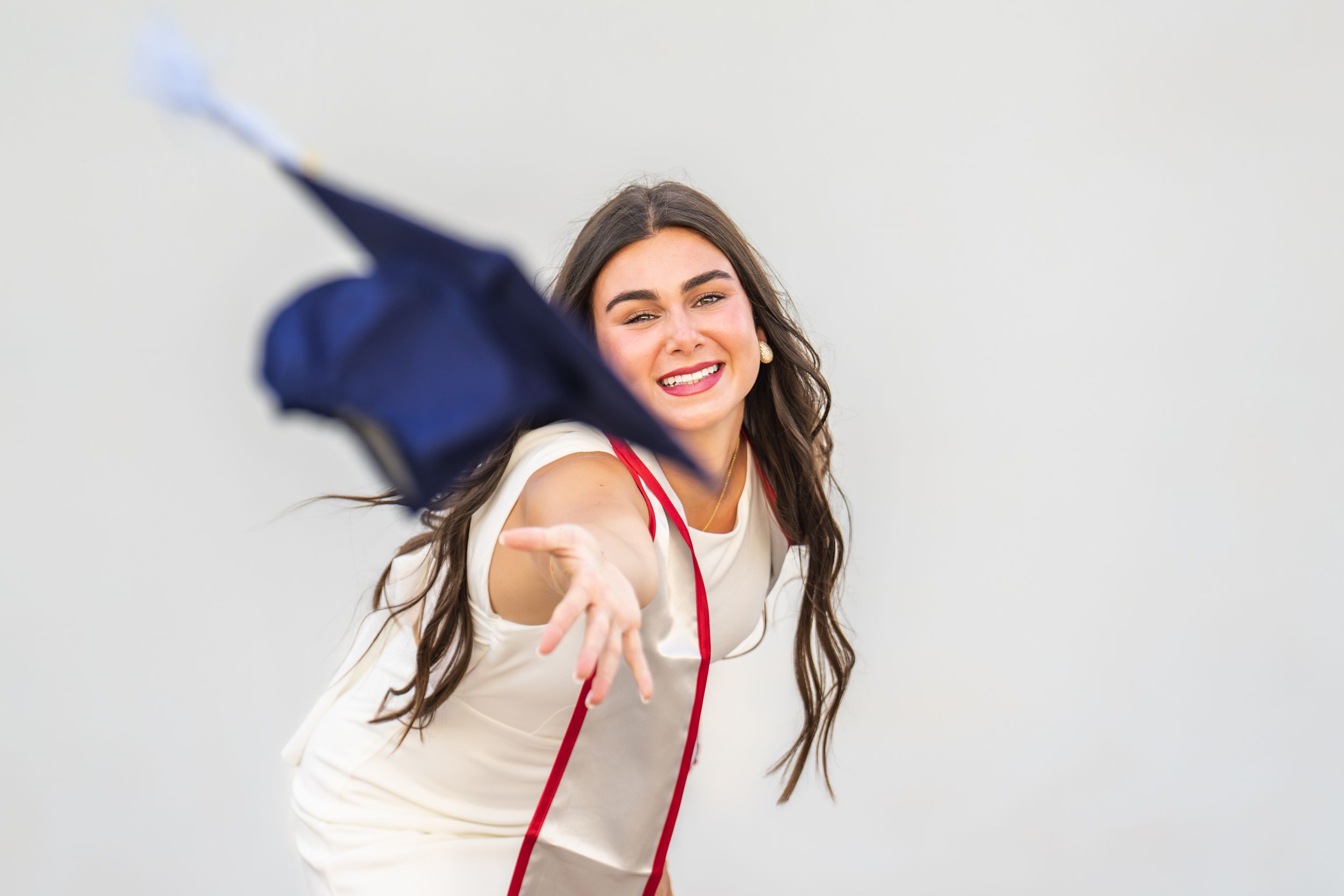 Smiling young woman in white top tossing graduation cap with red tassel against white background.