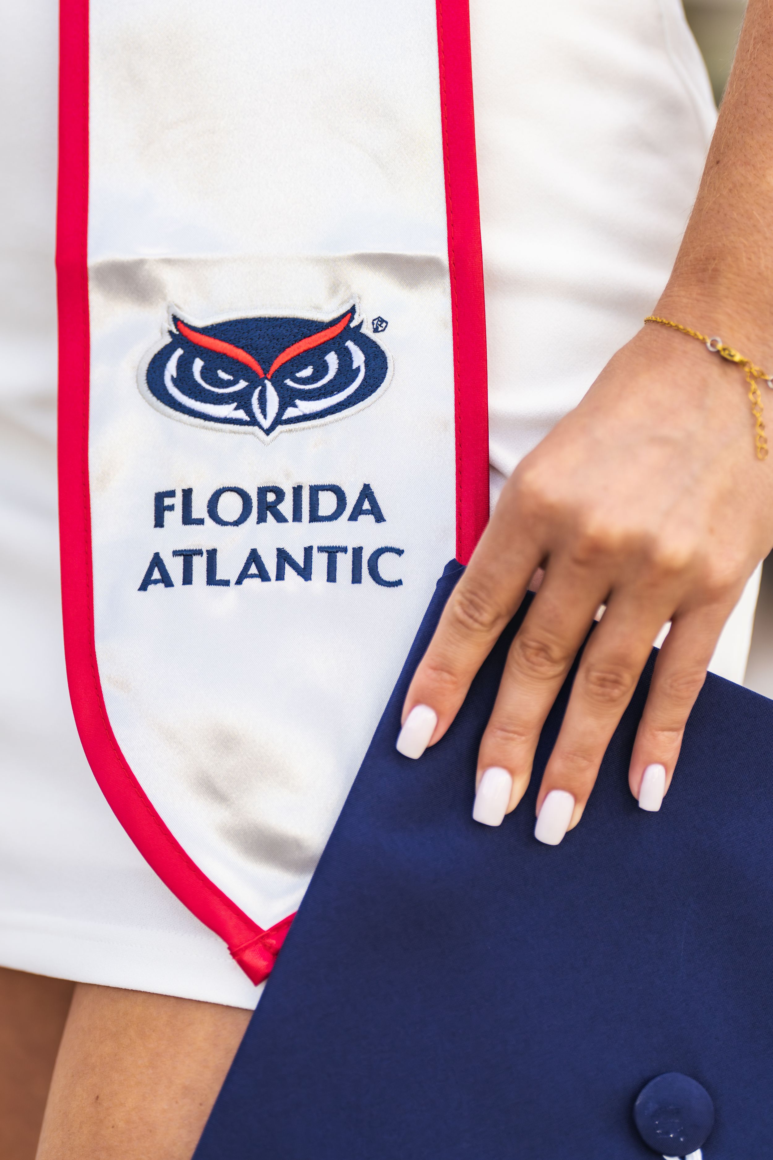 Florida Atlantic University graduation stole with owl mascot logo, held by a graduate wearing a gold bracelet.