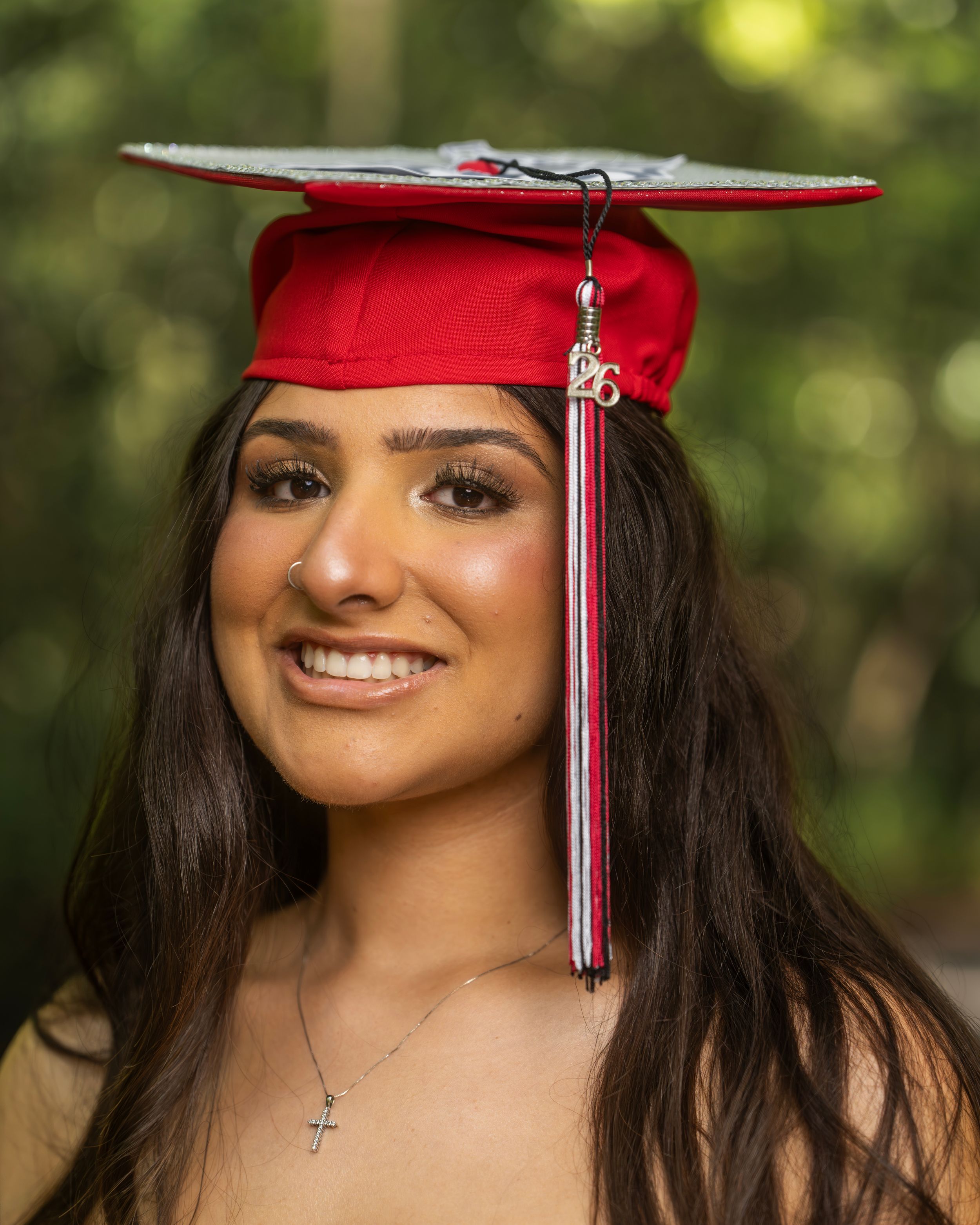 Young woman smiling in red graduation cap and gown with tassel, posing outdoors with green bokeh background.