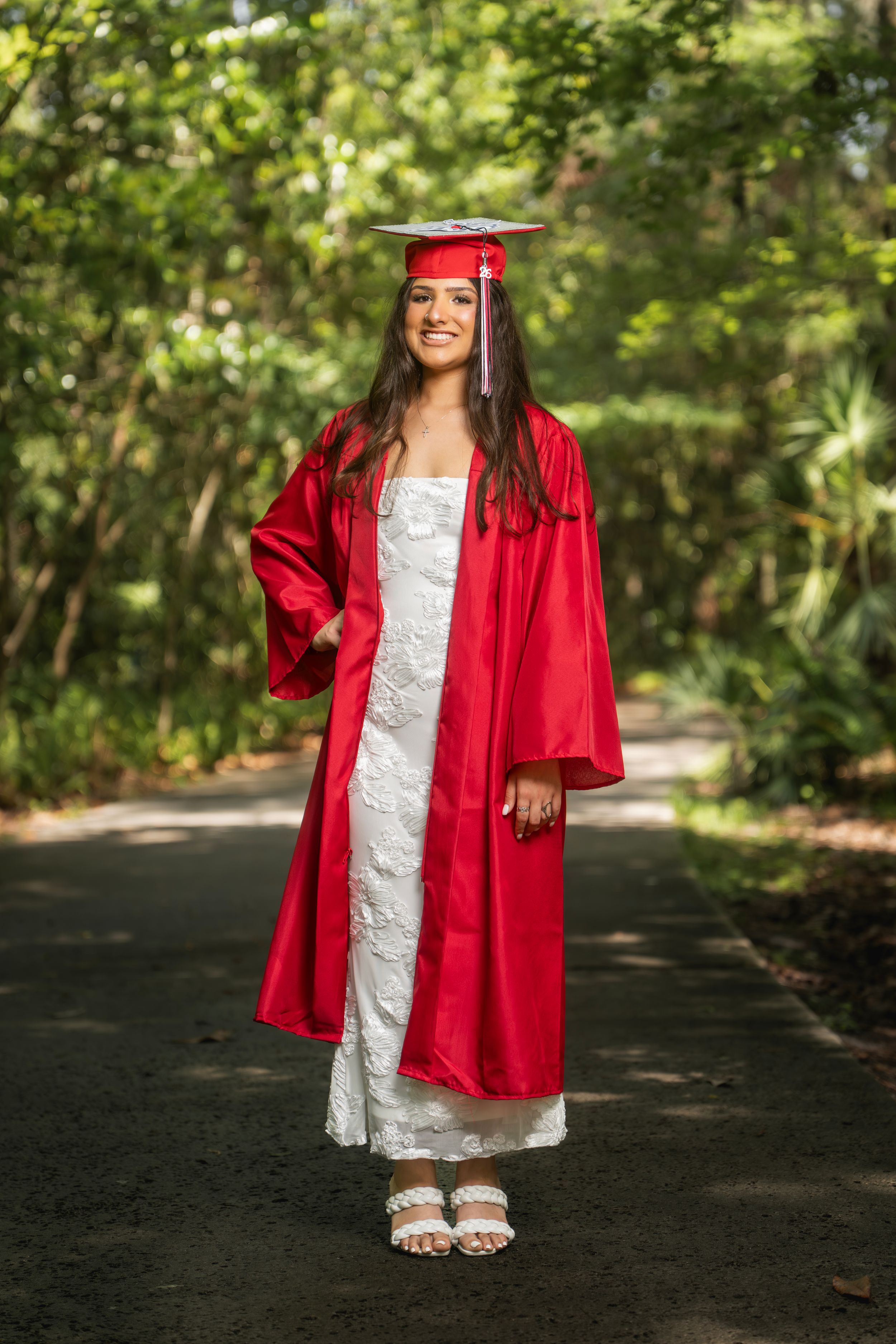 Young woman in red graduation cap and gown smiling outdoors on a tree-lined path wearing a white dress.