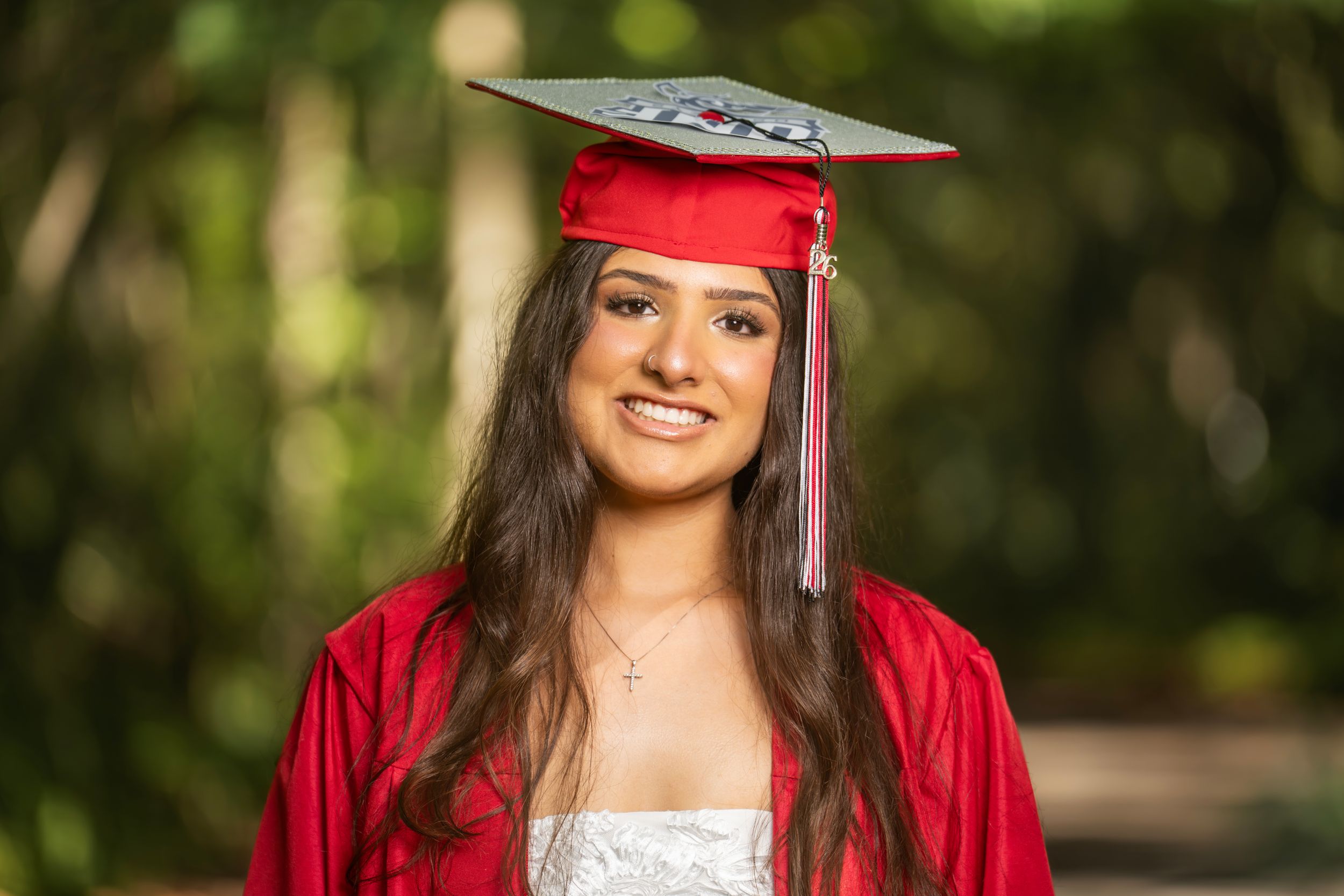 Young woman smiling in red graduation cap and gown outdoors with green trees in background.