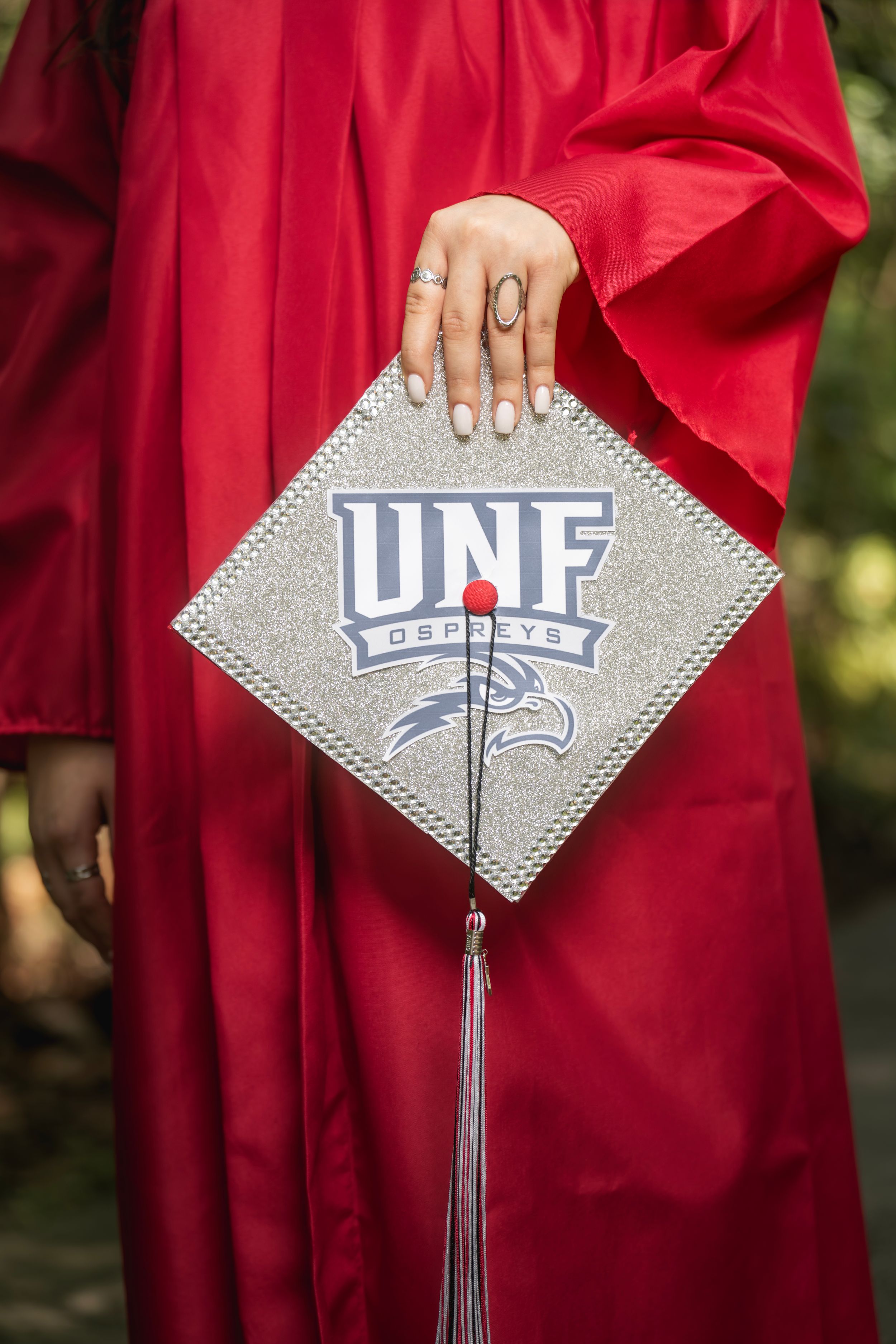 Close-up of UNF Ospreys decorated graduation cap held by graduate in red gown with glitter design.
