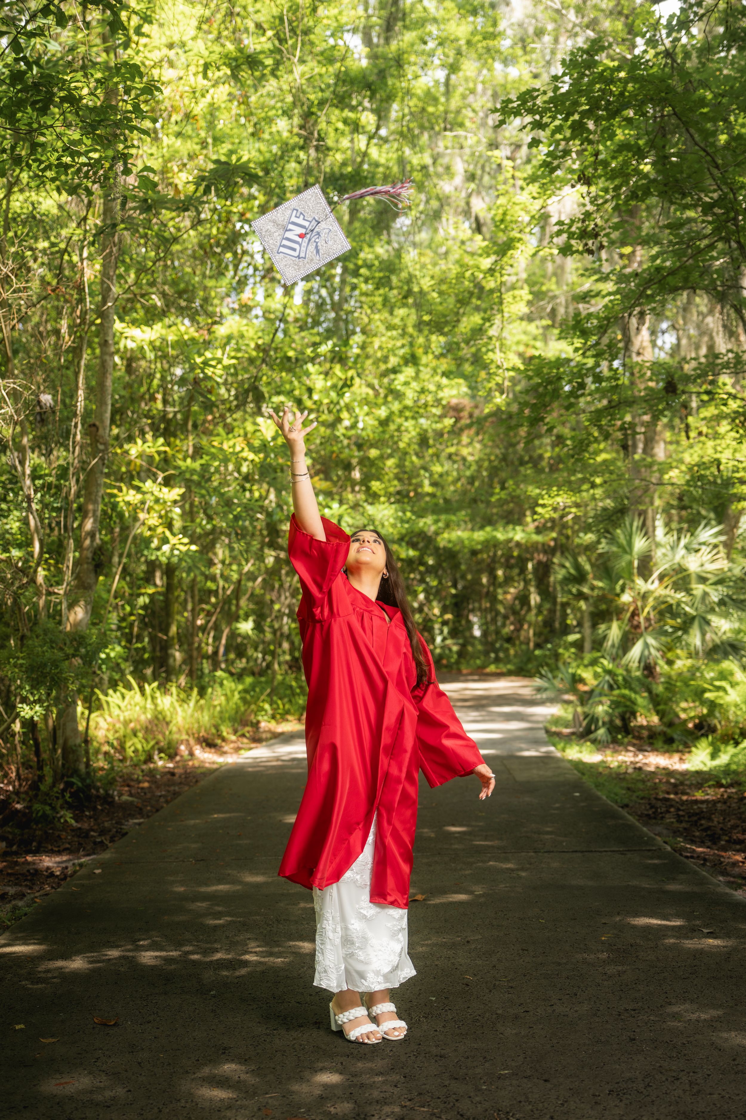 Excited graduate in red cap and gown tossing her graduation cap into the air on a scenic tree-lined path.