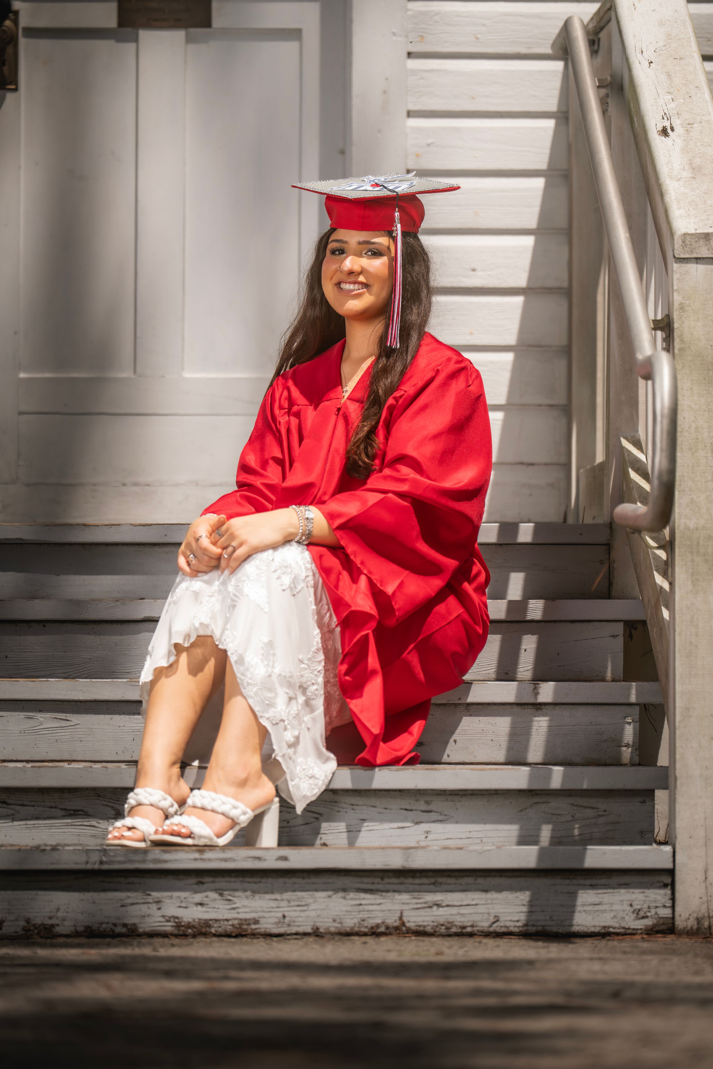 Young woman in red graduation cap and gown sitting on wooden steps smiling in bright sunlight.