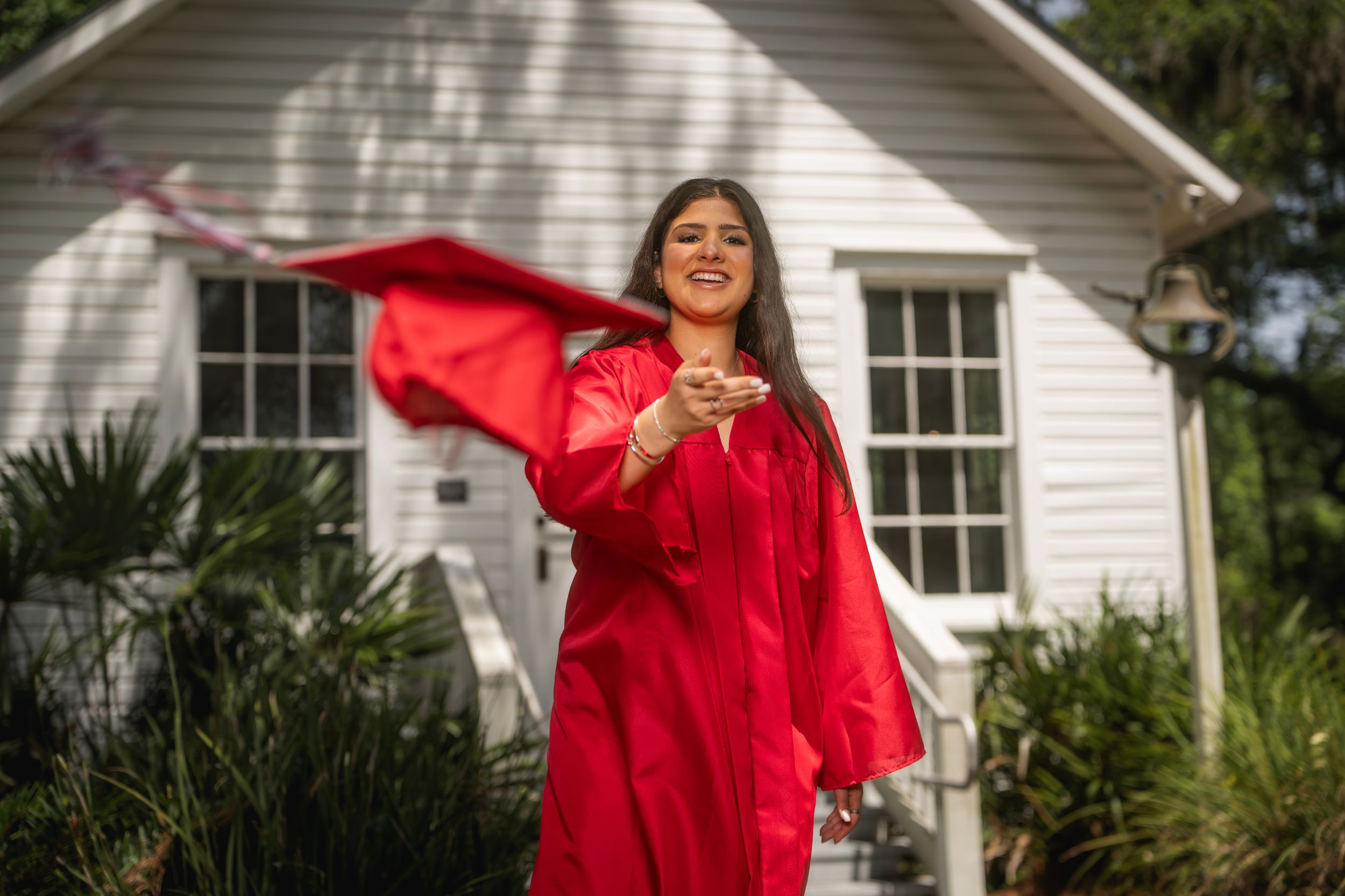 Young woman in red graduation gown and cap tosses her stole joyfully outside a white house.