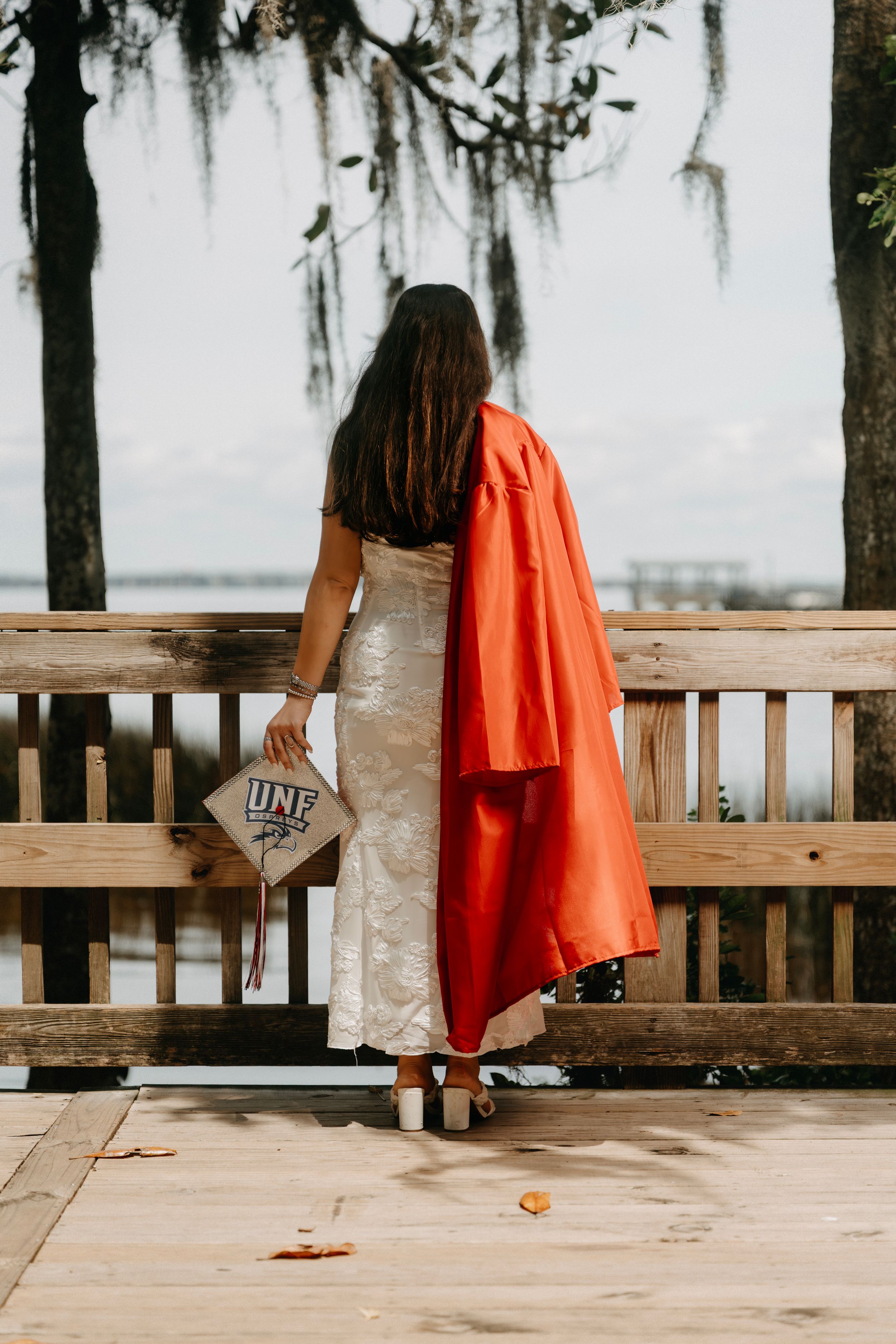 UNF graduate in red gown holding mortarboard cap while standing at a wooden waterfront railing.