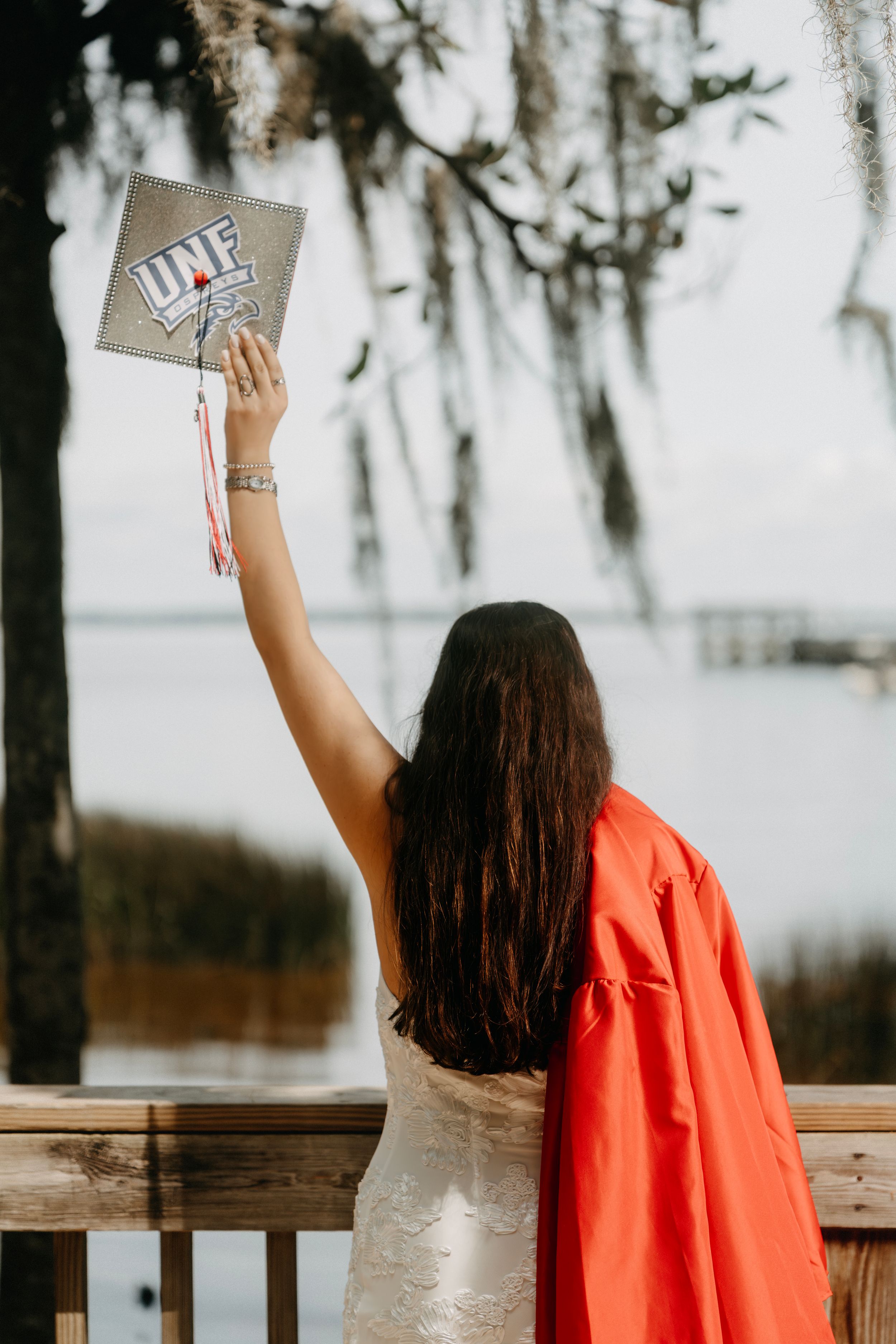 UNF graduate raises cap in celebration on waterfront deck wearing red gown and white dress.
