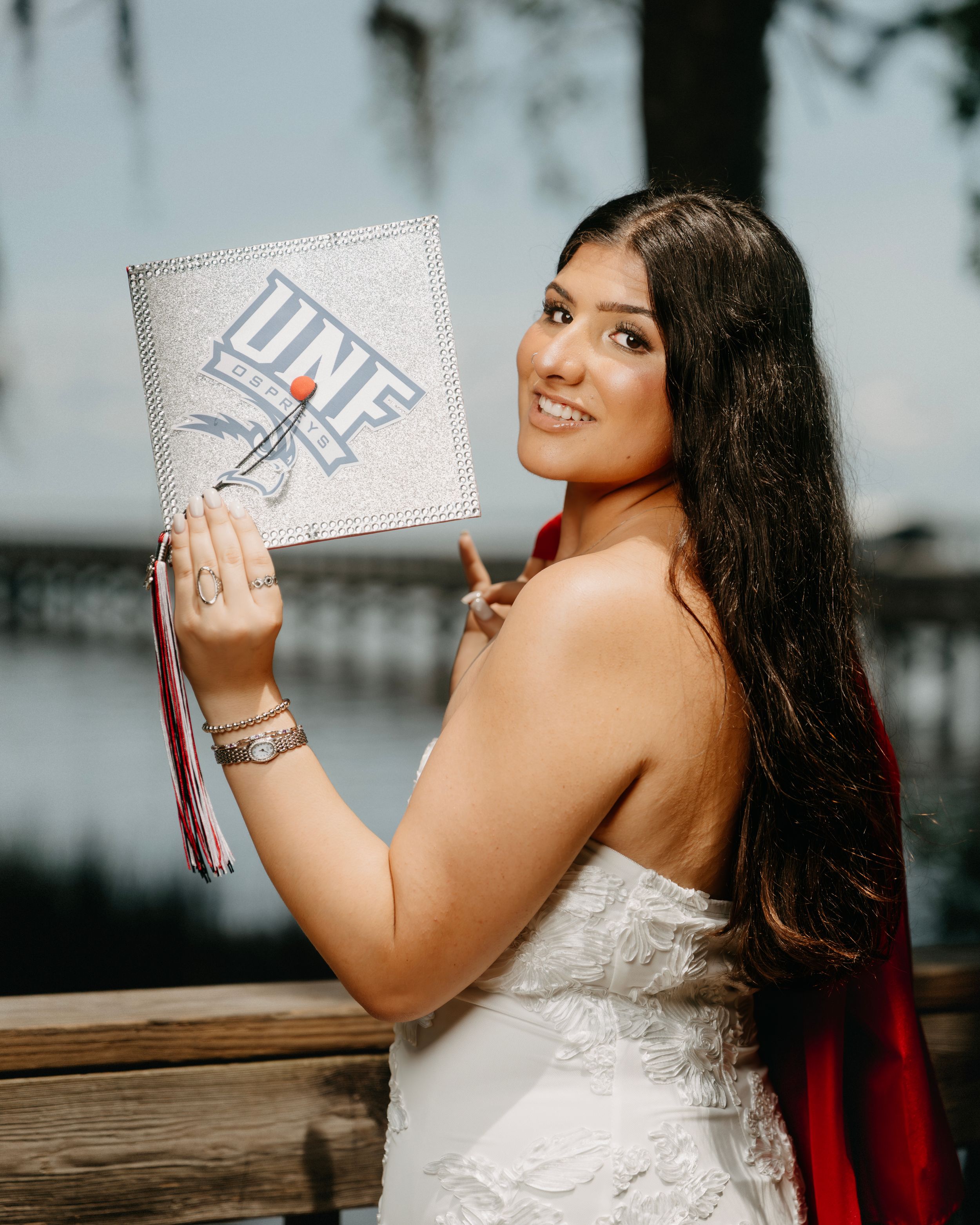 Smiling UNF graduate in white dress holding bedazzled mortarboard cap with red tassel outdoors.