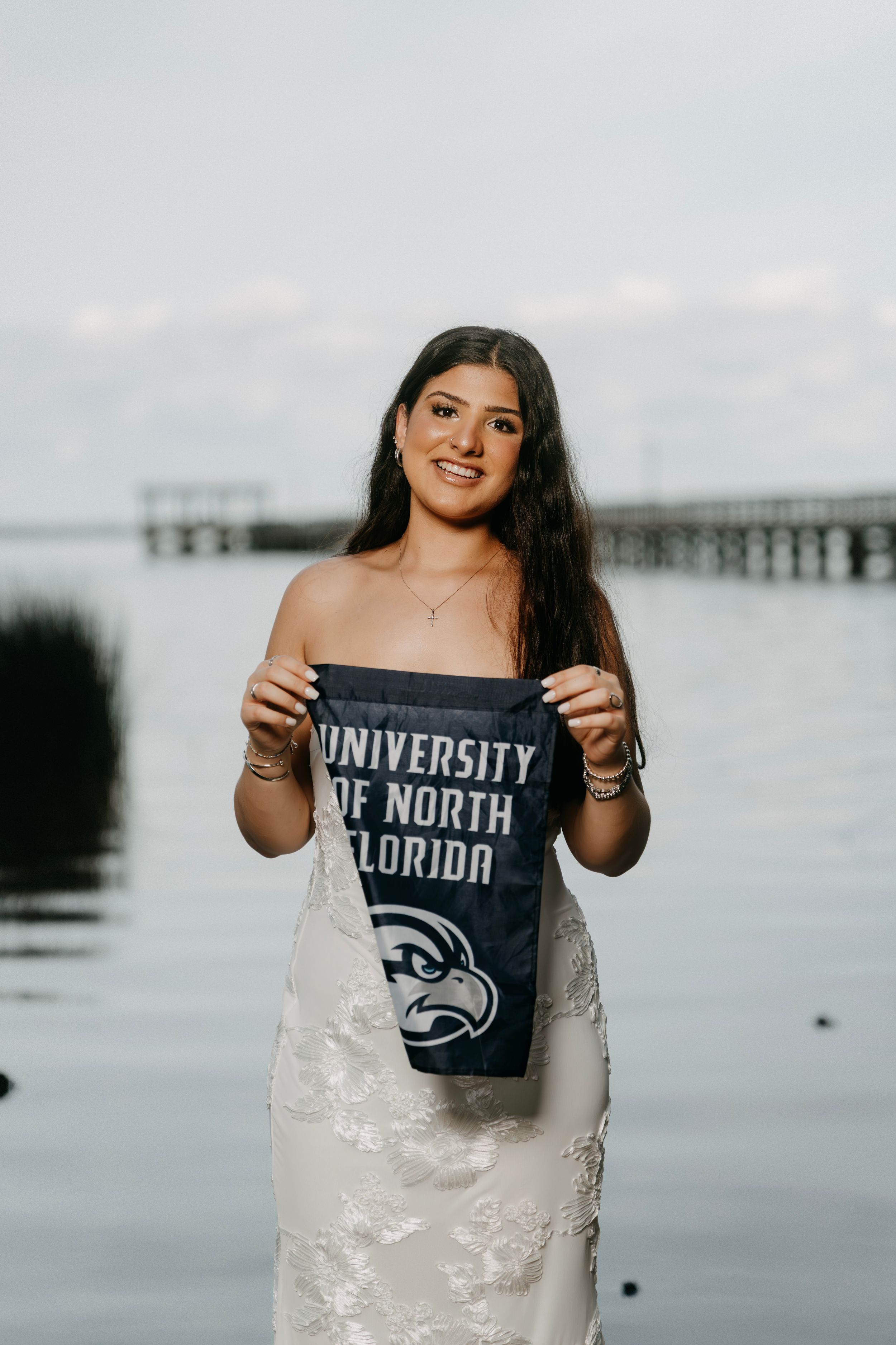 Smiling young woman in white dress holding University of North Florida pennant near waterfront.
