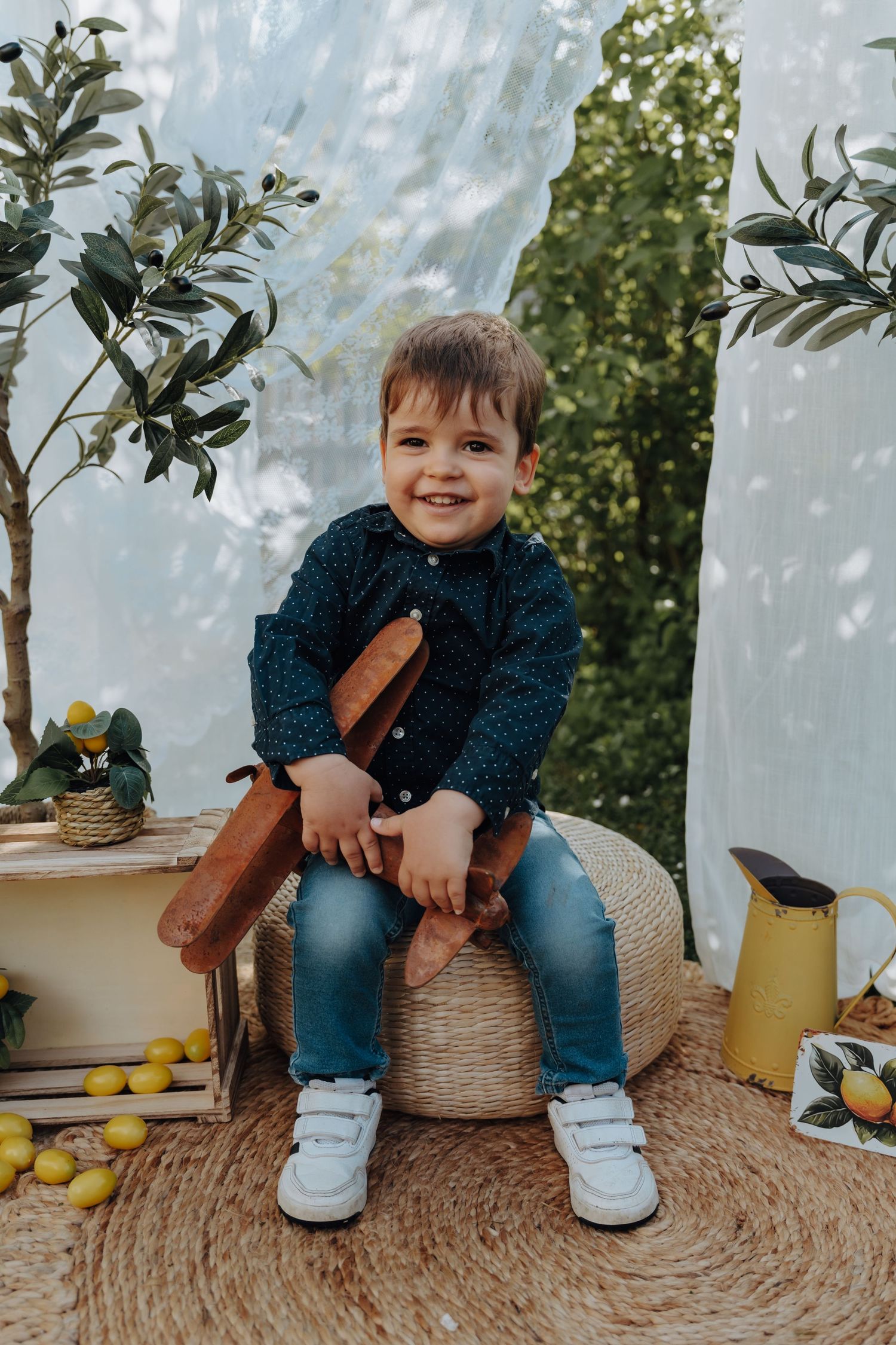 A smiling young boy in a dark shirt and jeans sits on a woven pouf surrounded by lemons and olive trees.