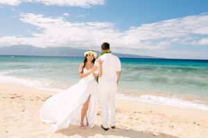 Bride and groom in white attire stand together on a Maui beach with stunning turquoise ocean views.