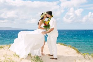Bride and groom kiss on sandy dunes during their tropical Hawaiian beach wedding with vibrant bouquet.