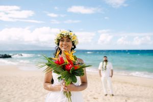 Smiling Hawaiian bride wearing floral crown holds colorful tropical bouquet on a sunny Maui beach.