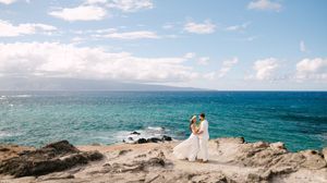 Bride and groom share a romantic moment on rocky coastal cliffs overlooking turquoise Hawaiian waters.