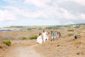 Wedding party walks along a scenic dirt path through golden Hawaiian landscape under a cloudy sky.