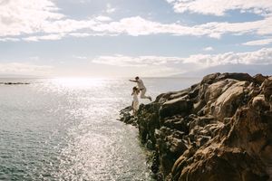Couple jumping between rocky cliffs by the ocean on a sunny day.