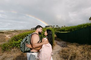 Couple embracing on a sandy path with a rainbow in the cloudy sky behind them.