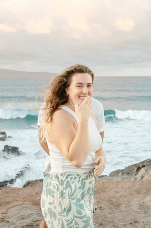 A woman laughs joyfully on a rocky Hawaiian beach with ocean waves crashing behind her.