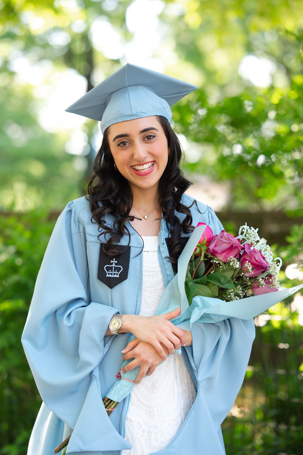 Smiling young woman in light blue graduation cap and gown holding pink flowers bouquet outdoors.