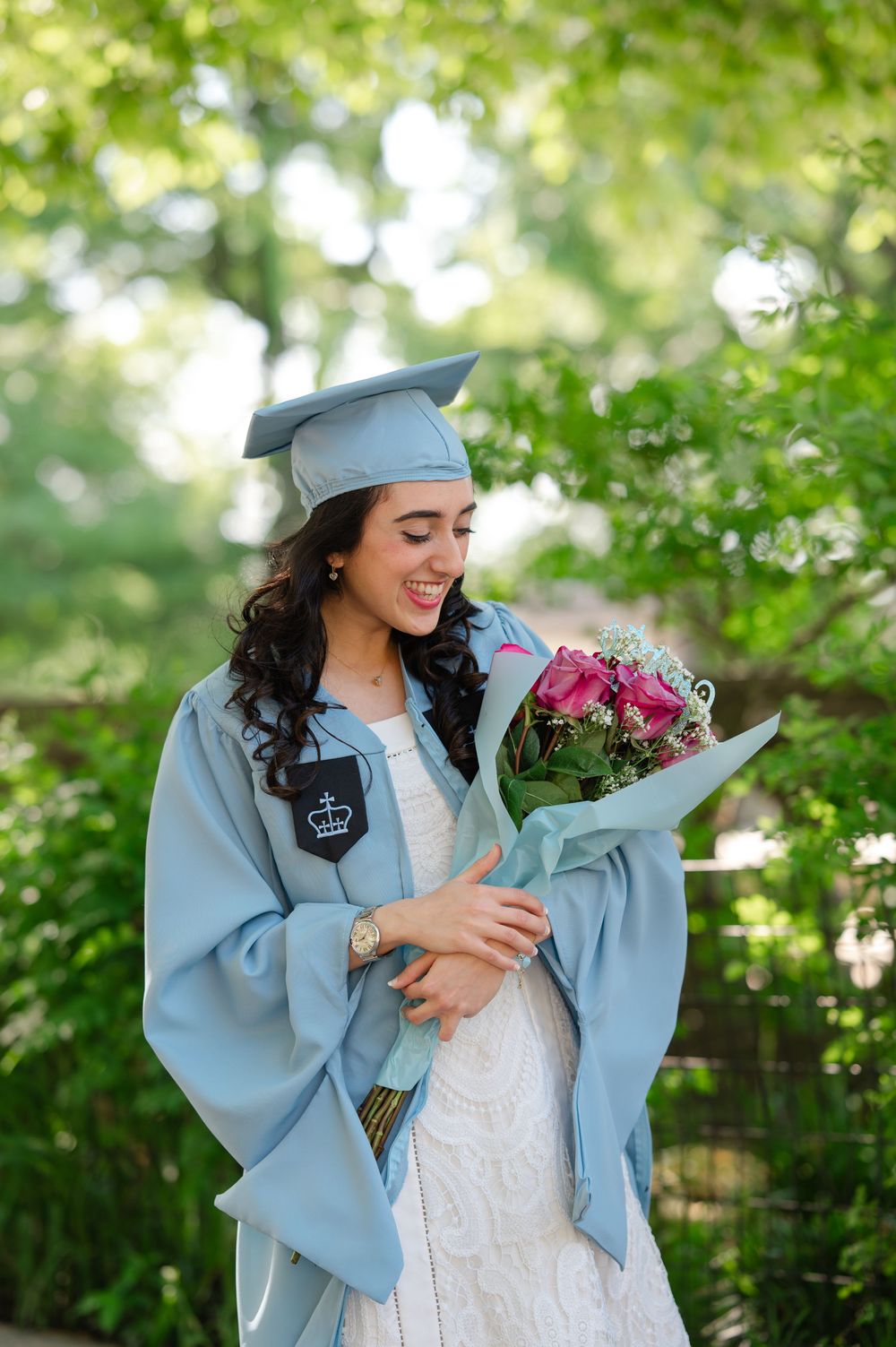 Smiling female graduate in light blue cap and gown holding pink flower bouquet outdoors.