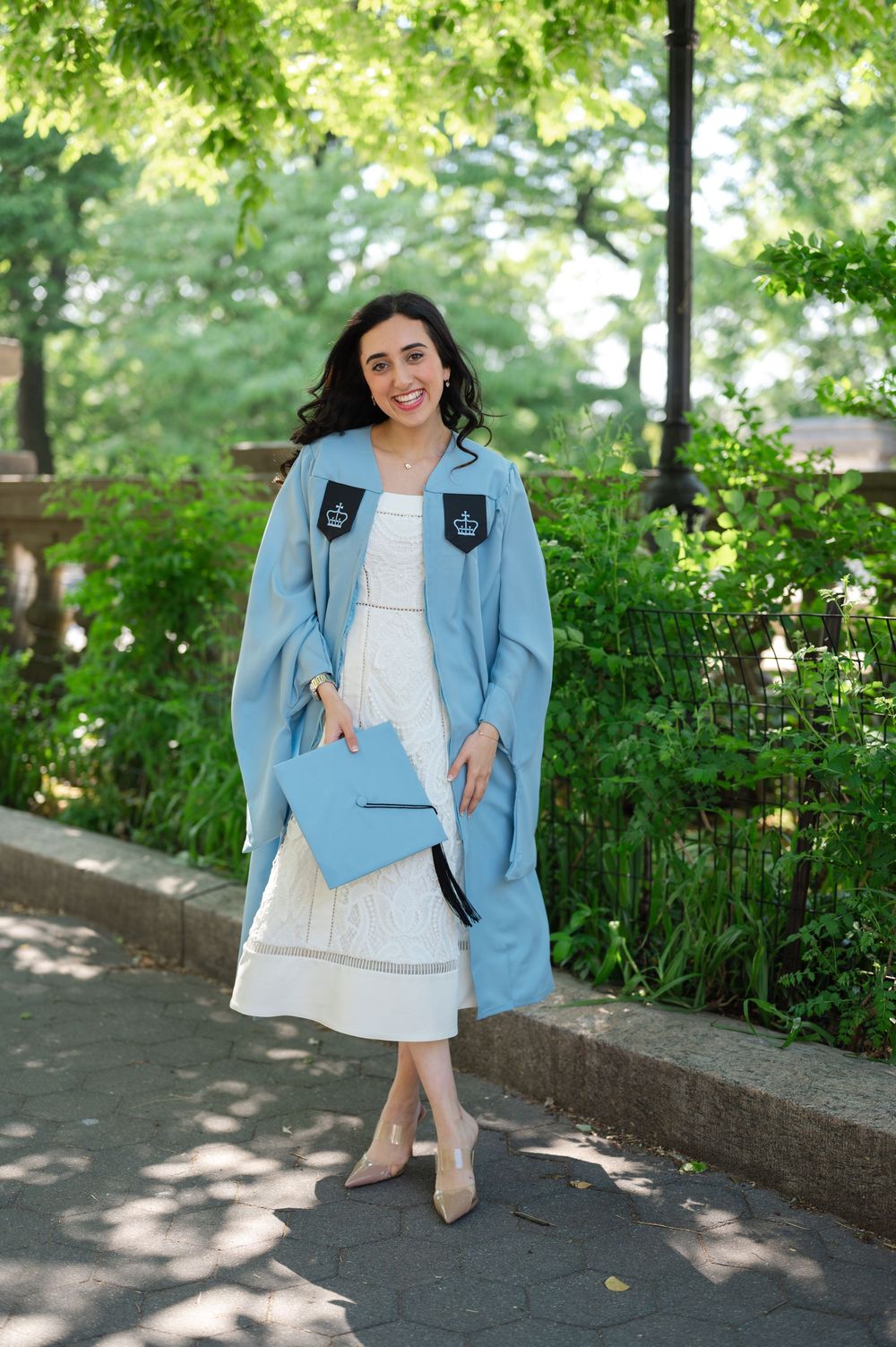 Smiling graduate in light blue cap and gown holding diploma outdoors in lush green park setting.