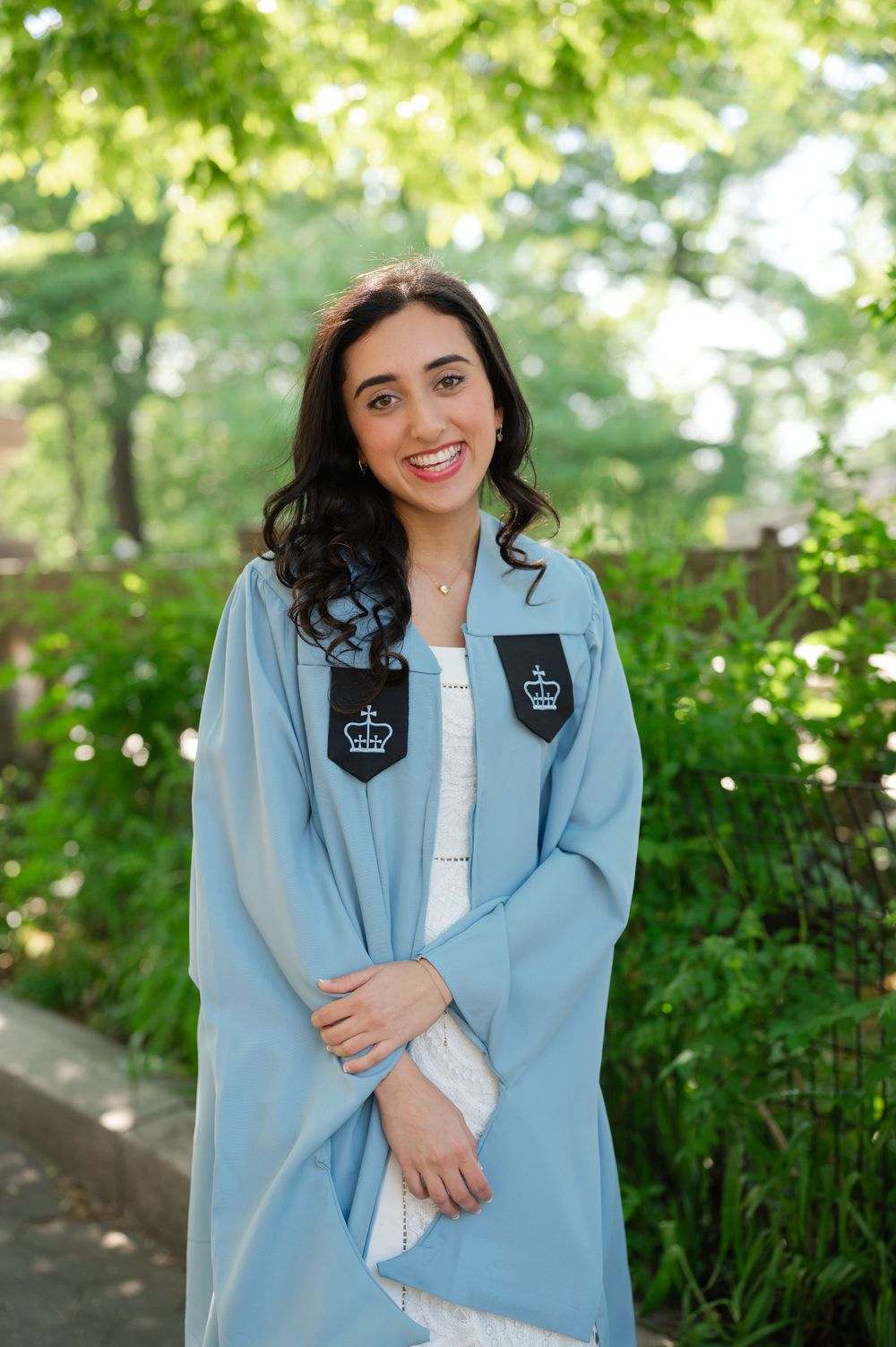 Smiling young woman in light blue Columbia University graduation gown posing outdoors among lush green trees.