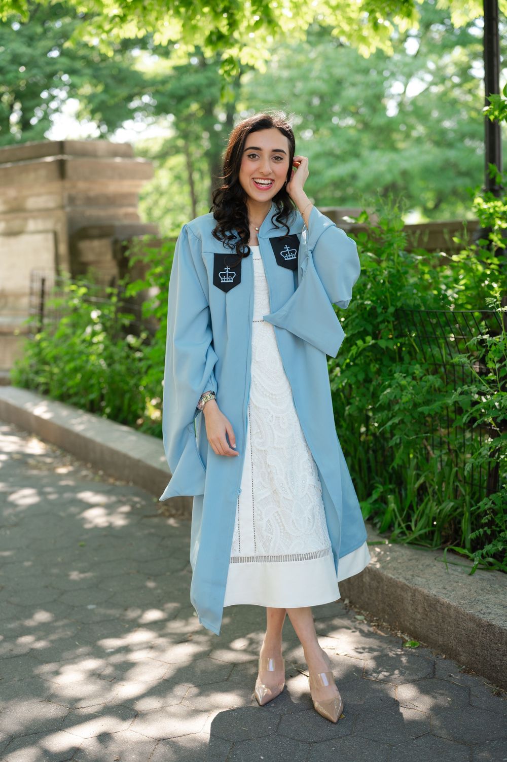 Smiling woman in light blue graduation gown over white dress, posing outdoors in a lush green park setting.