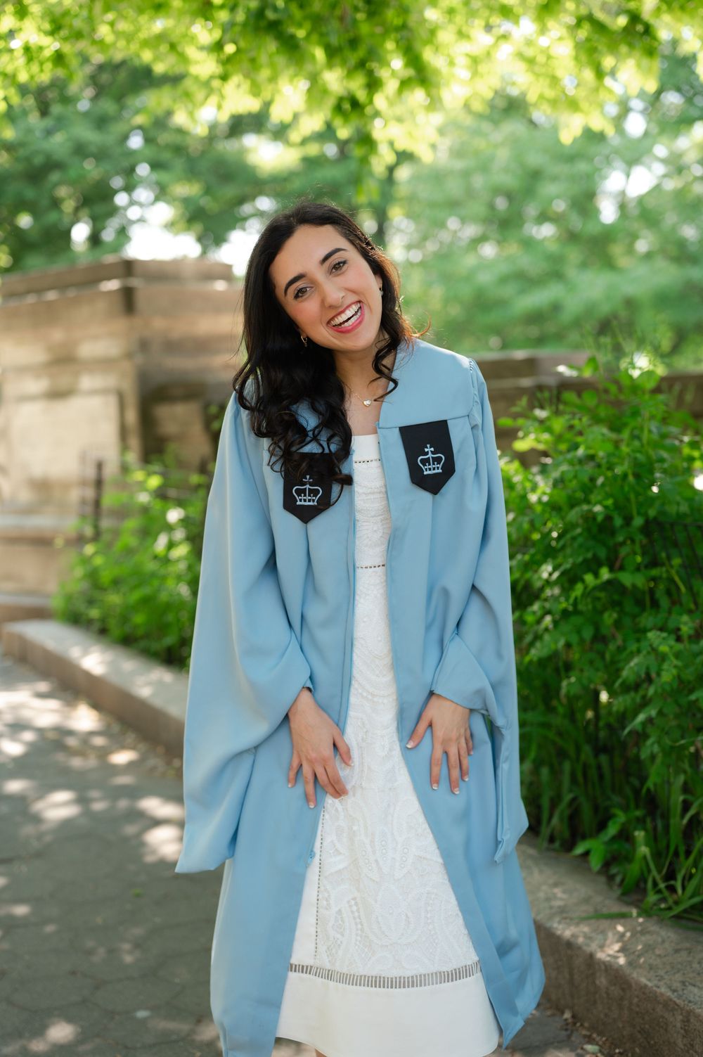 Smiling young woman in light blue Columbia University graduation gown over white lace dress, posing outdoors.