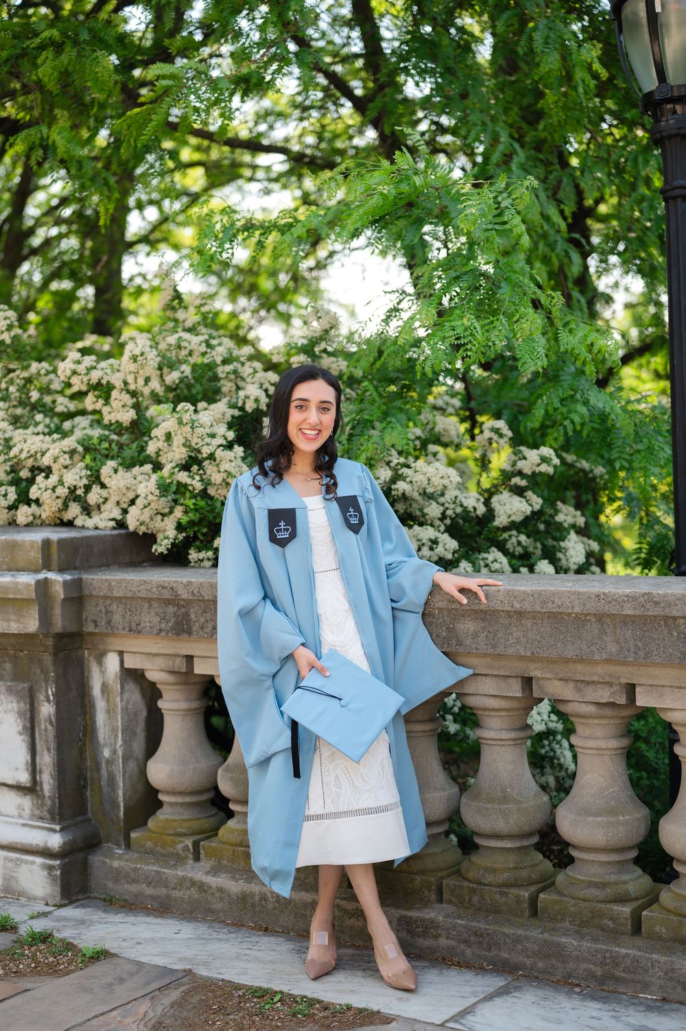 Smiling graduate in light blue graduation gown holding cap beside stone balustrade with white flowering trees.