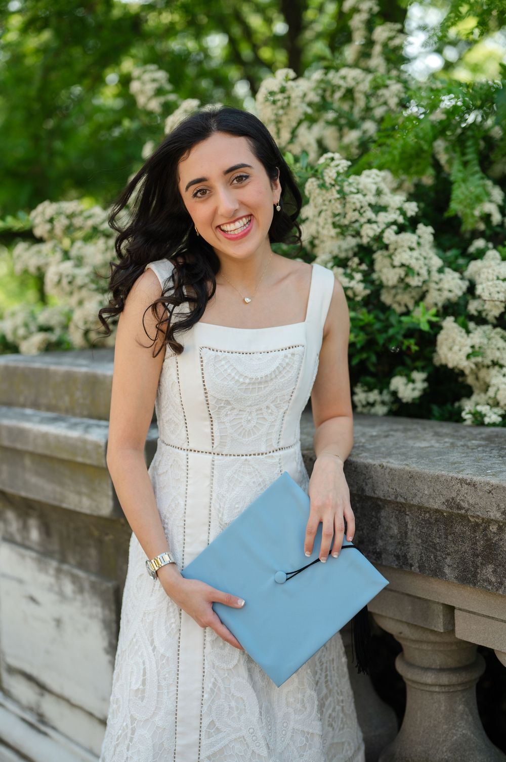 Young woman in white lace dress holding light blue graduation cap, smiling outdoors near blooming white flowers.