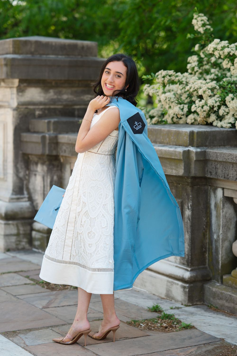 Young woman in white dress holding light blue graduation gown over shoulder, smiling outdoors near stone balustrade.