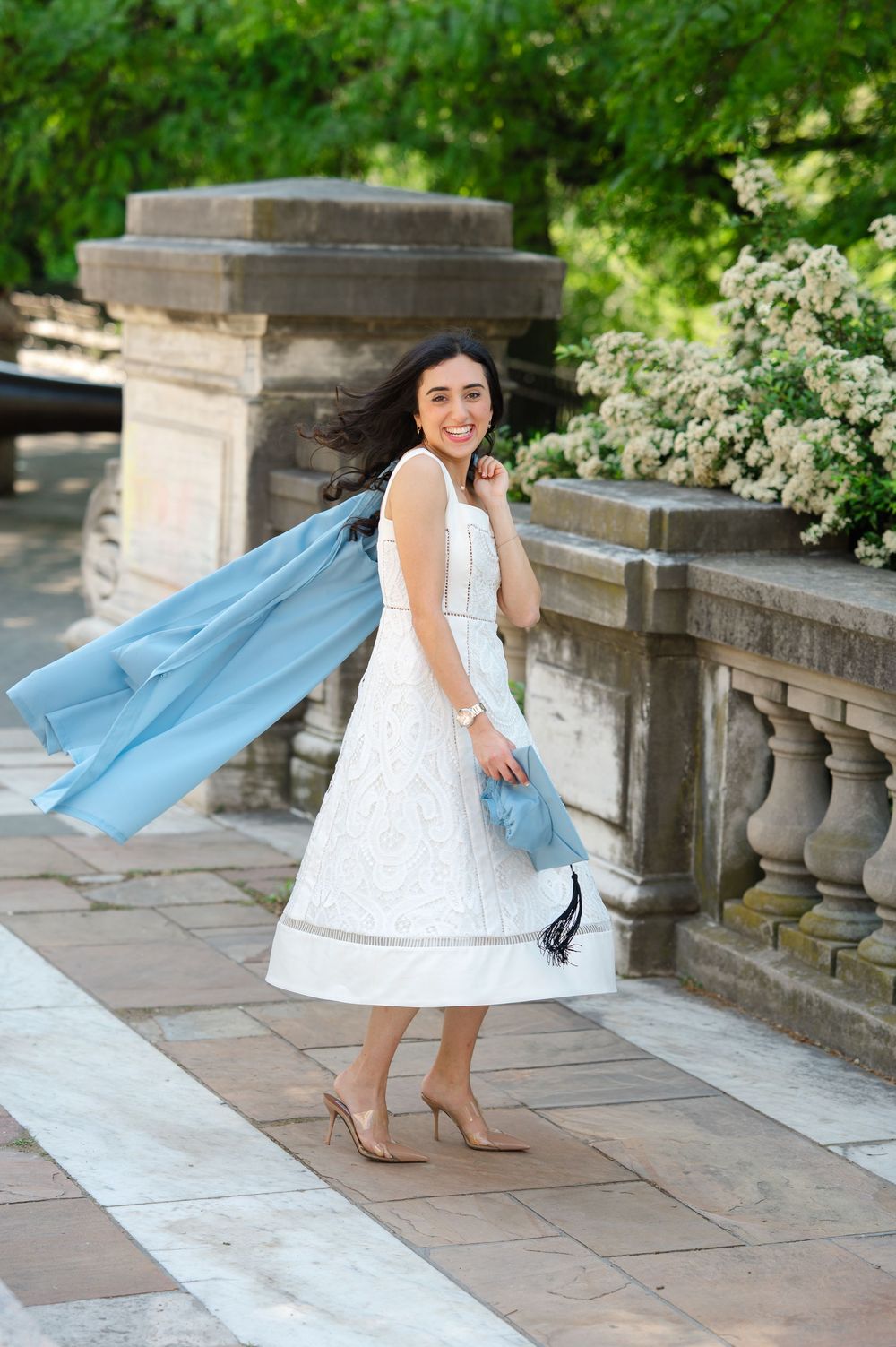 Woman in white midi dress holding light blue coat and clutch, smiling outdoors near stone balustrade with flowers.