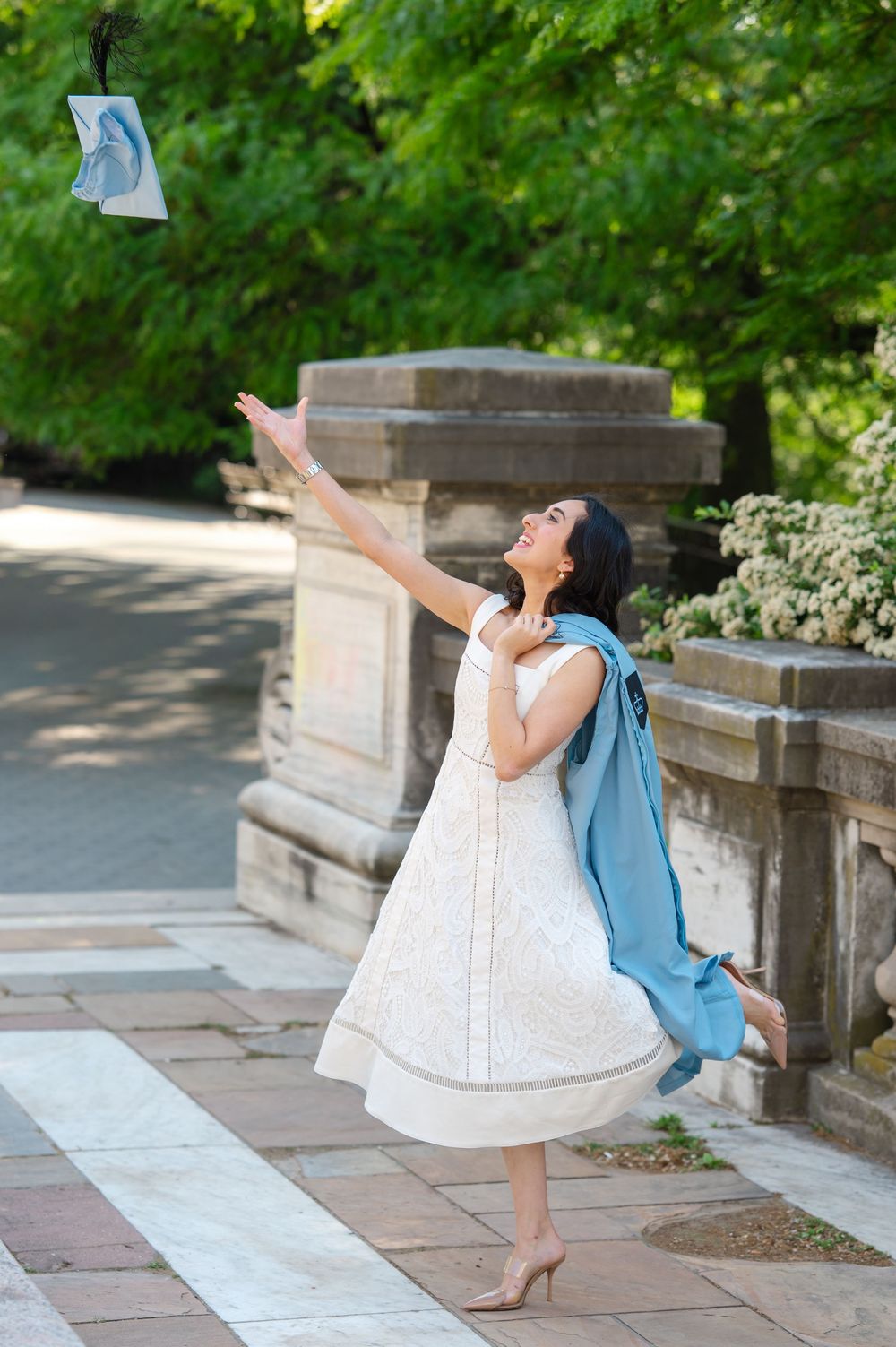Woman in white dress tossing graduation cap outdoors, wearing blue academic hood, celebrating commencement.