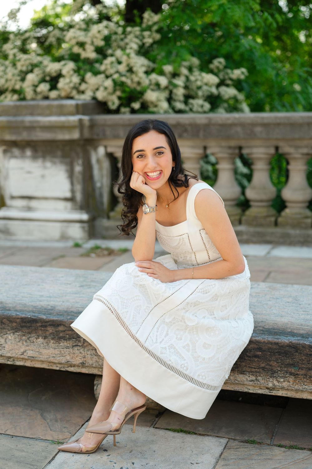 Young woman in white dress sitting on stone steps in a garden with blooming flowers.