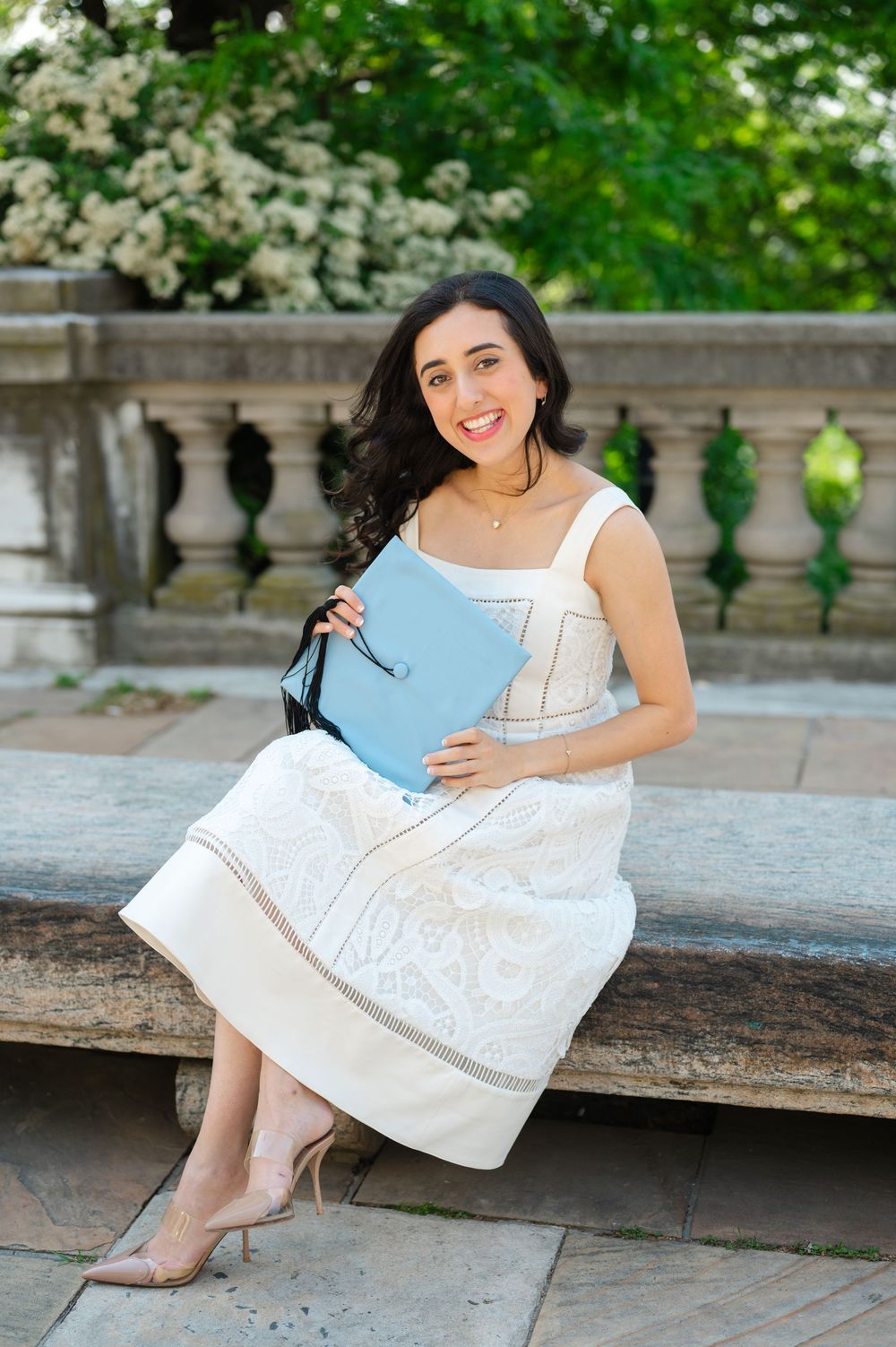 Happy graduate in white lace dress holding light blue diploma folder, seated on stone steps outdoors.