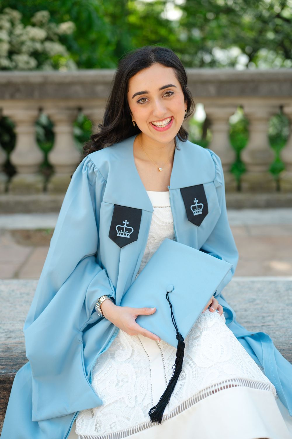 Joyful young woman in Columbia University light blue graduation gown holding mortarboard cap, seated outdoors.