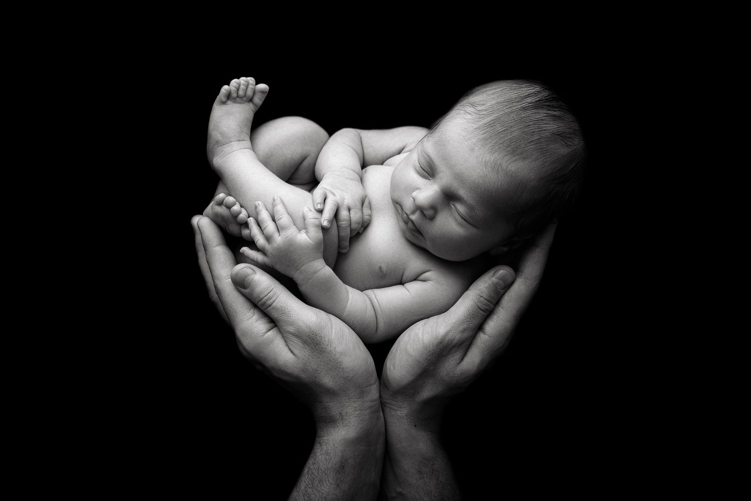 Black and white photo of a newborn baby cradled gently in adult hands against a dark background.