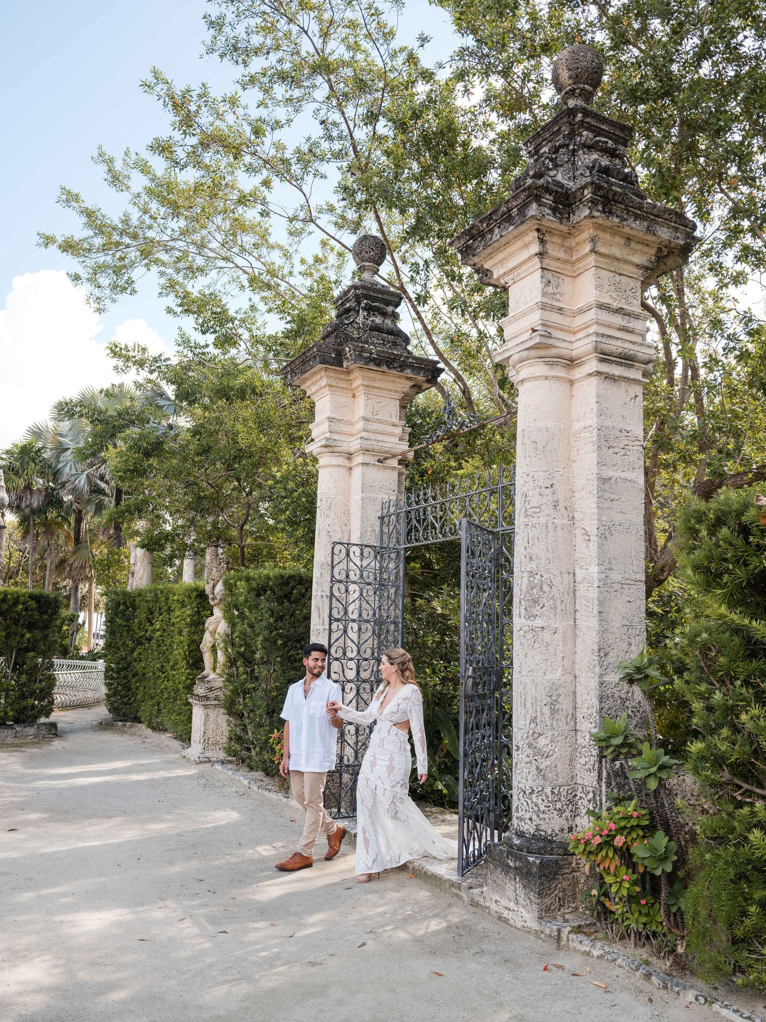 Bride and groom walk through an ornate garden gate at Vizcaya Gardens, surrounded by historic stone pillars and tropical greenery.