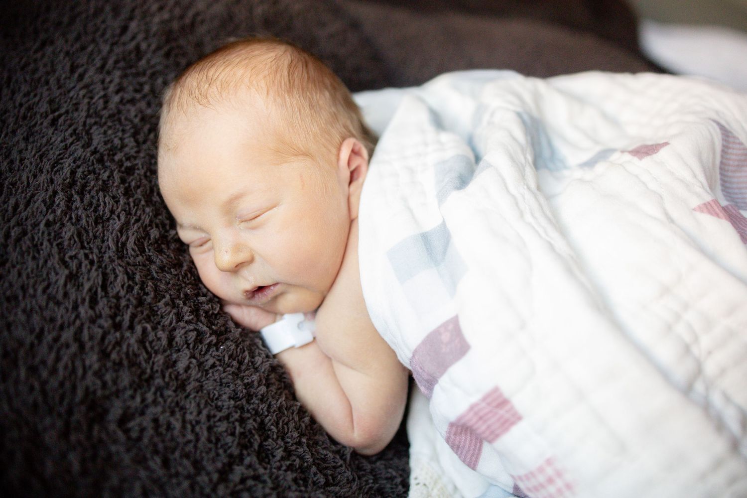Peaceful newborn baby sleeping wrapped in a soft muslin swaddle blanket on a dark fuzzy surface.