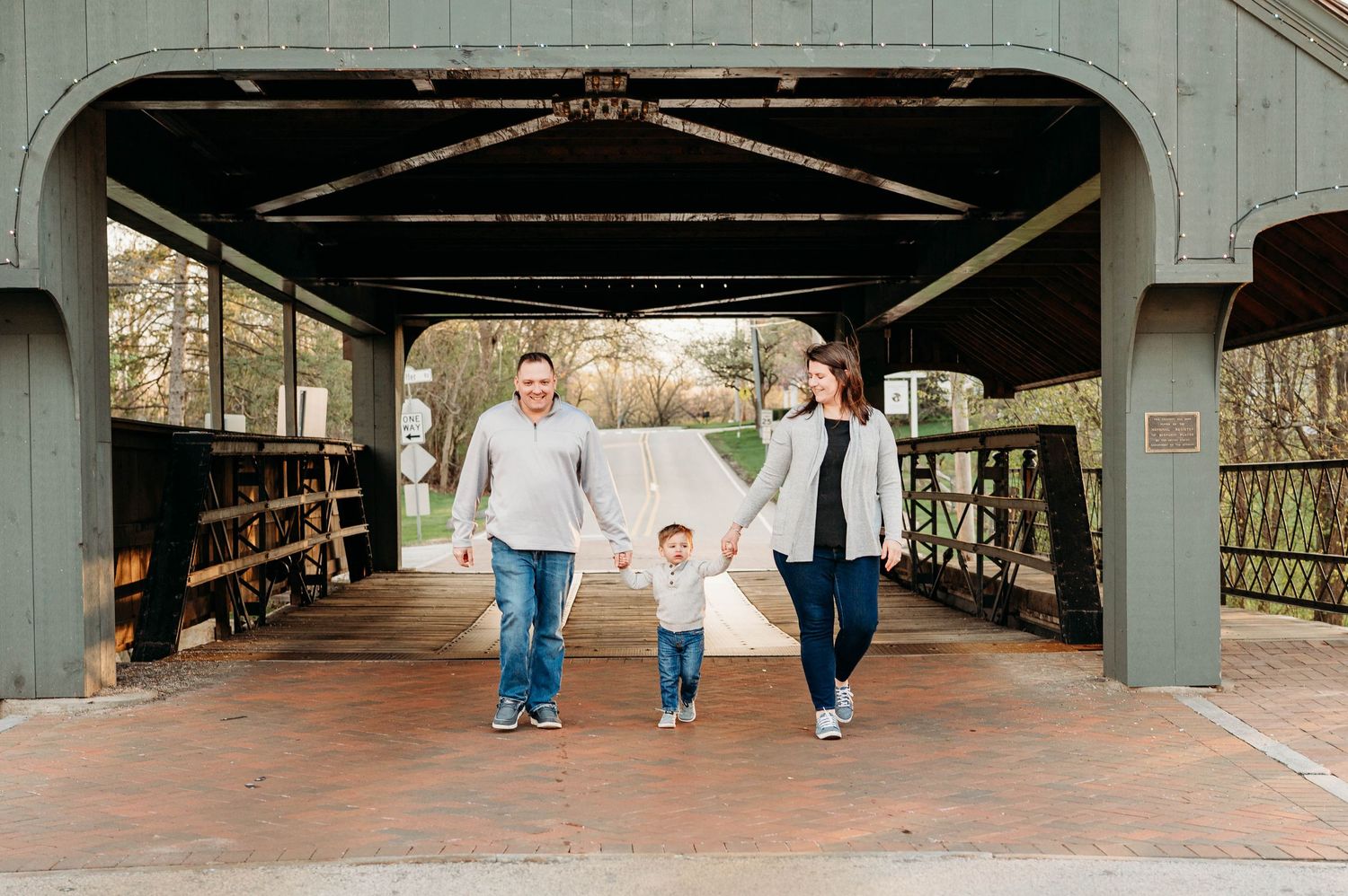 Family of three holding hands walking through a covered bridge outdoors during a fall family photo session.