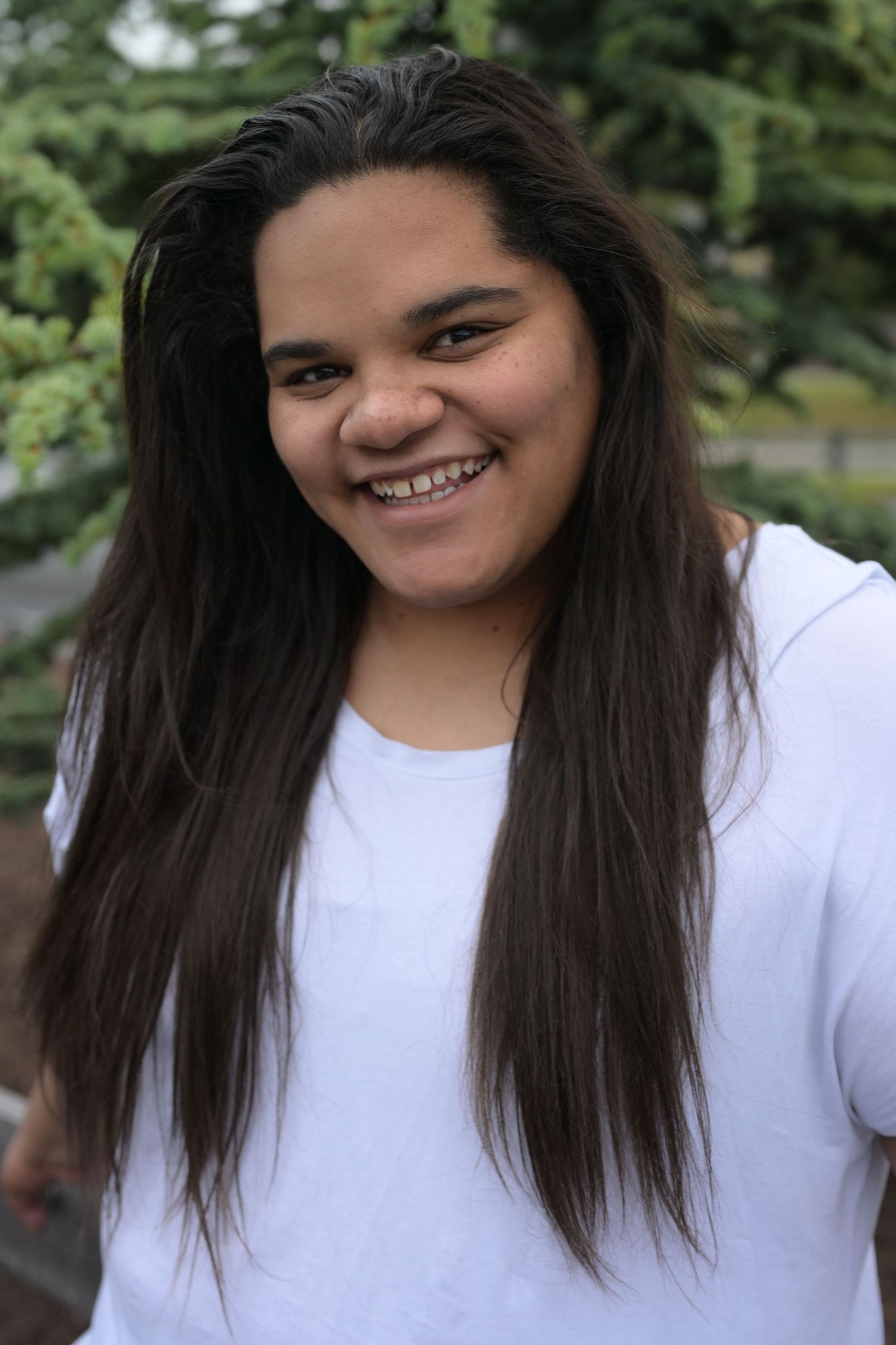 Smiling young woman with long dark hair wearing a white t-shirt outdoors with green trees in background.