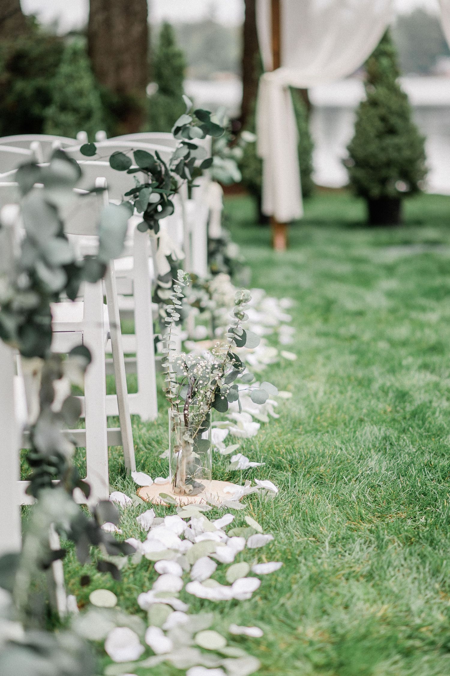 Outdoor wedding aisle with white chairs, eucalyptus garlands, glass vases, and white petals on lush green grass.