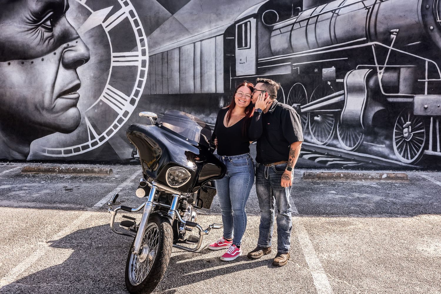 Couple sharing a kiss beside a motorcycle in front of a colorful urban mural.