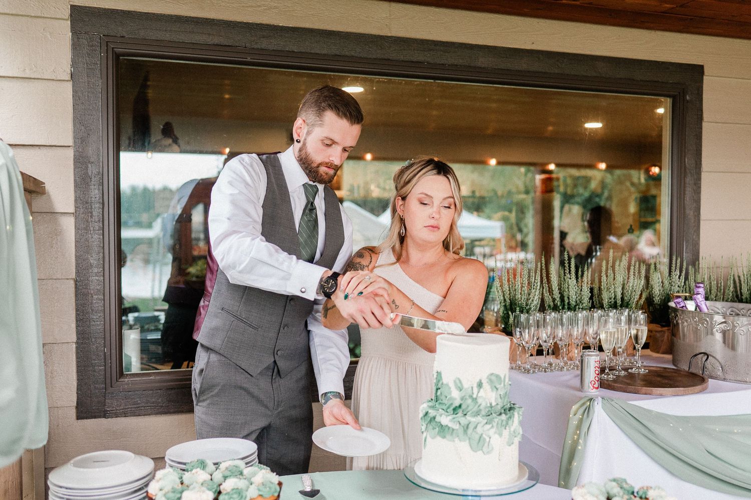Bride and groom cutting their white wedding cake with greenery decor at an outdoor lakeside reception.