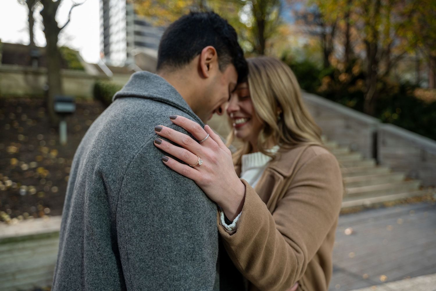 Romantic couple embracing outdoors in autumn, woman in tan coat smiling, man in gray coat, urban park setting.