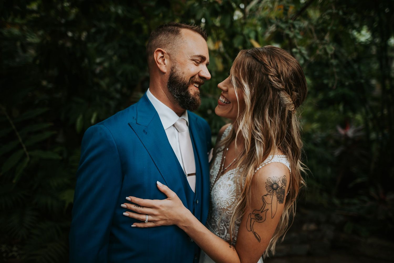 A smiling bride and groom share a romantic moment outdoors, surrounded by lush green foliage on their wedding day.