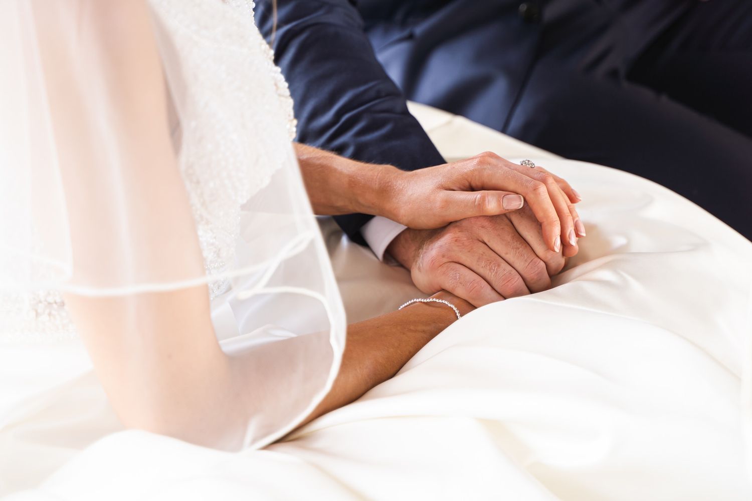 Bride and groom holding hands tenderly during wedding ceremony, close-up of couple in white dress and navy suit.